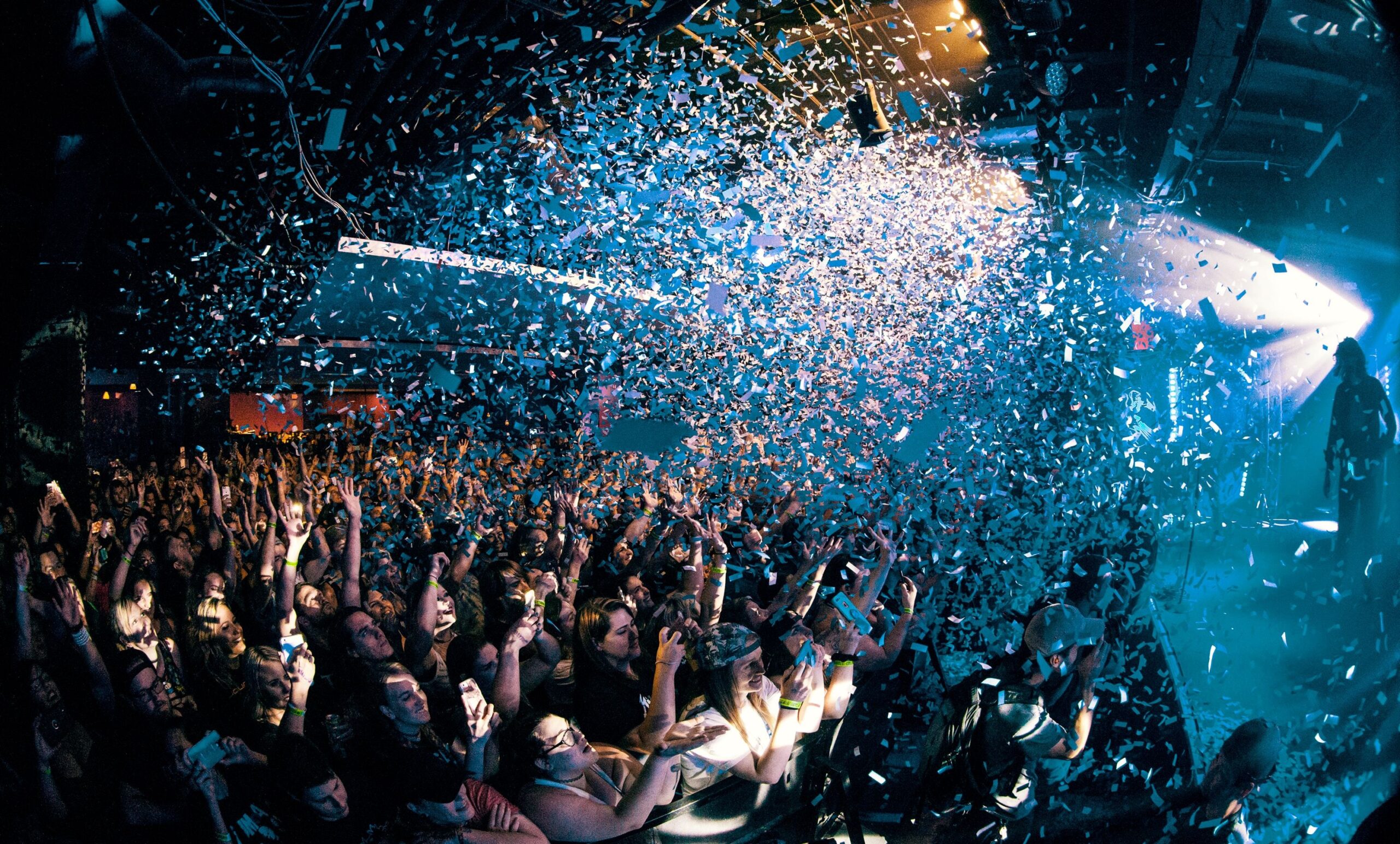 A lively concert scene where a crowd of fans raises their hands and phones in excitement as a storm of blue and white confetti rains down from above. Bright stage lights illuminate the venue, creating an energetic and festive atmosphere.