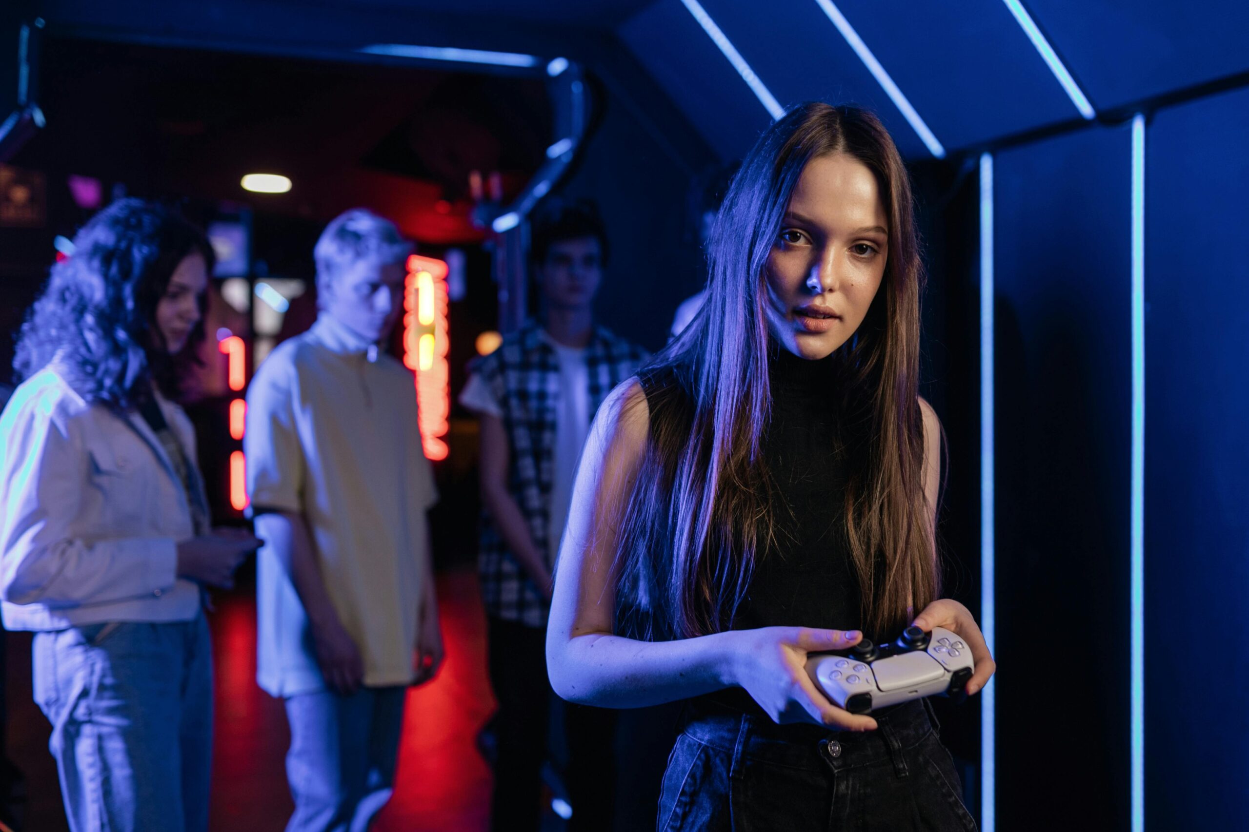 A young woman holding a gaming controller stands in the foreground of a futuristic arcade with neon blue and red lighting. She looks directly at the camera with a focused expression. In the background, a group of friends watches, their faces softly illuminated by the arcade's colorful glow.
