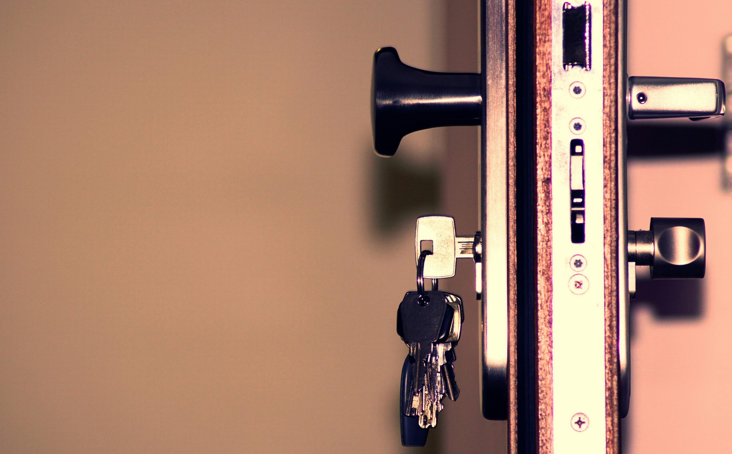 A close-up of a door lock with a key inserted. The key is attached to a ring holding multiple other keys. The door's metal frame and locking mechanism are visible, with a black door handle in the background. The warm lighting highlights the metallic surface, emphasizing the secure and sturdy structure.