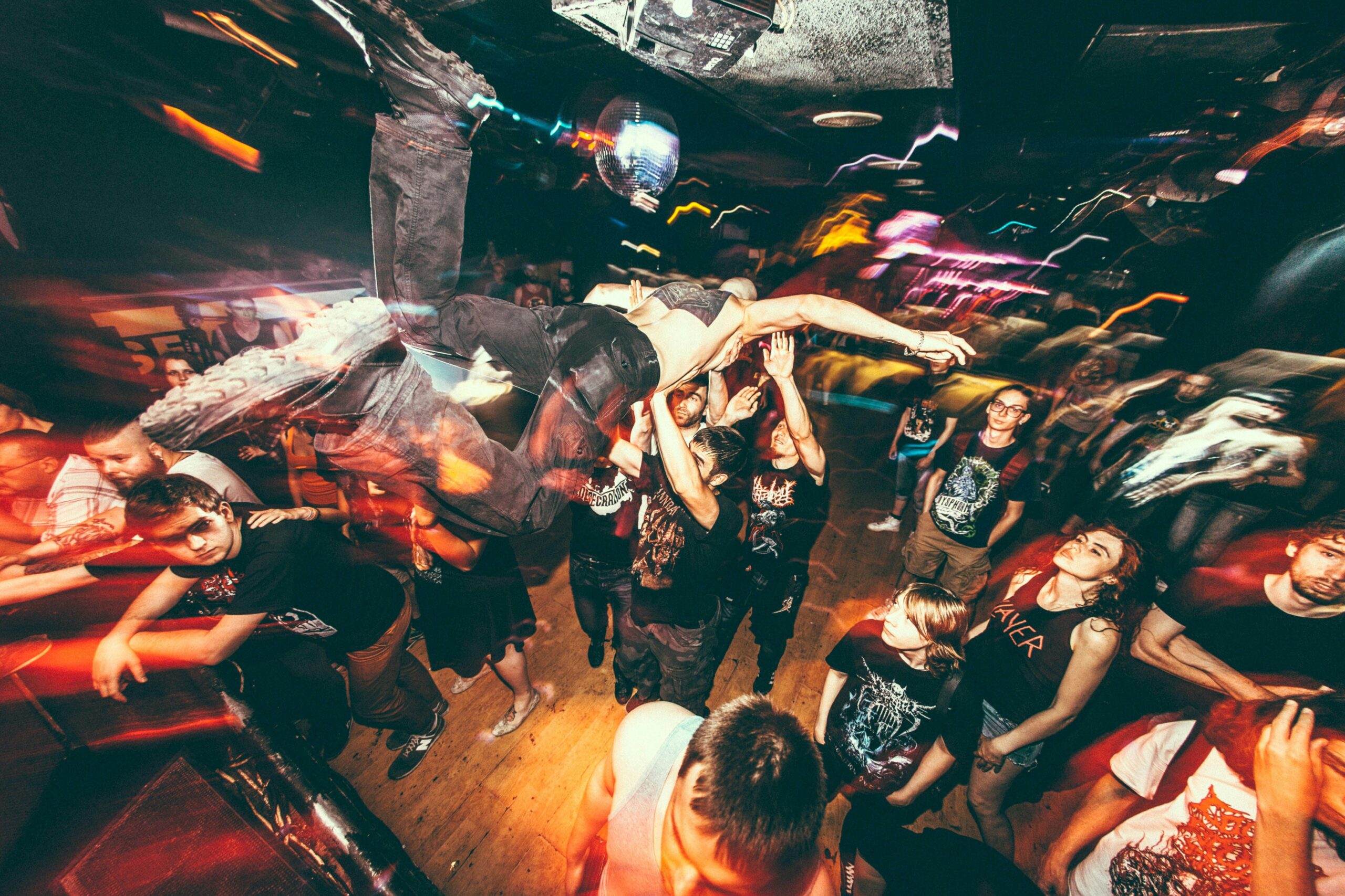 A chaotic and energetic concert scene featuring a crowd surfer being carried across a group of fans. The crowd reaches upward, supporting the person mid-air, while colorful, blurred stage lighting and motion effects add a sense of dynamic movement and intensity. The atmosphere is lively, with attendees immersed in the music and experience. Several people in the foreground wear band-themed shirts, embodying the raw energy of a live music event.