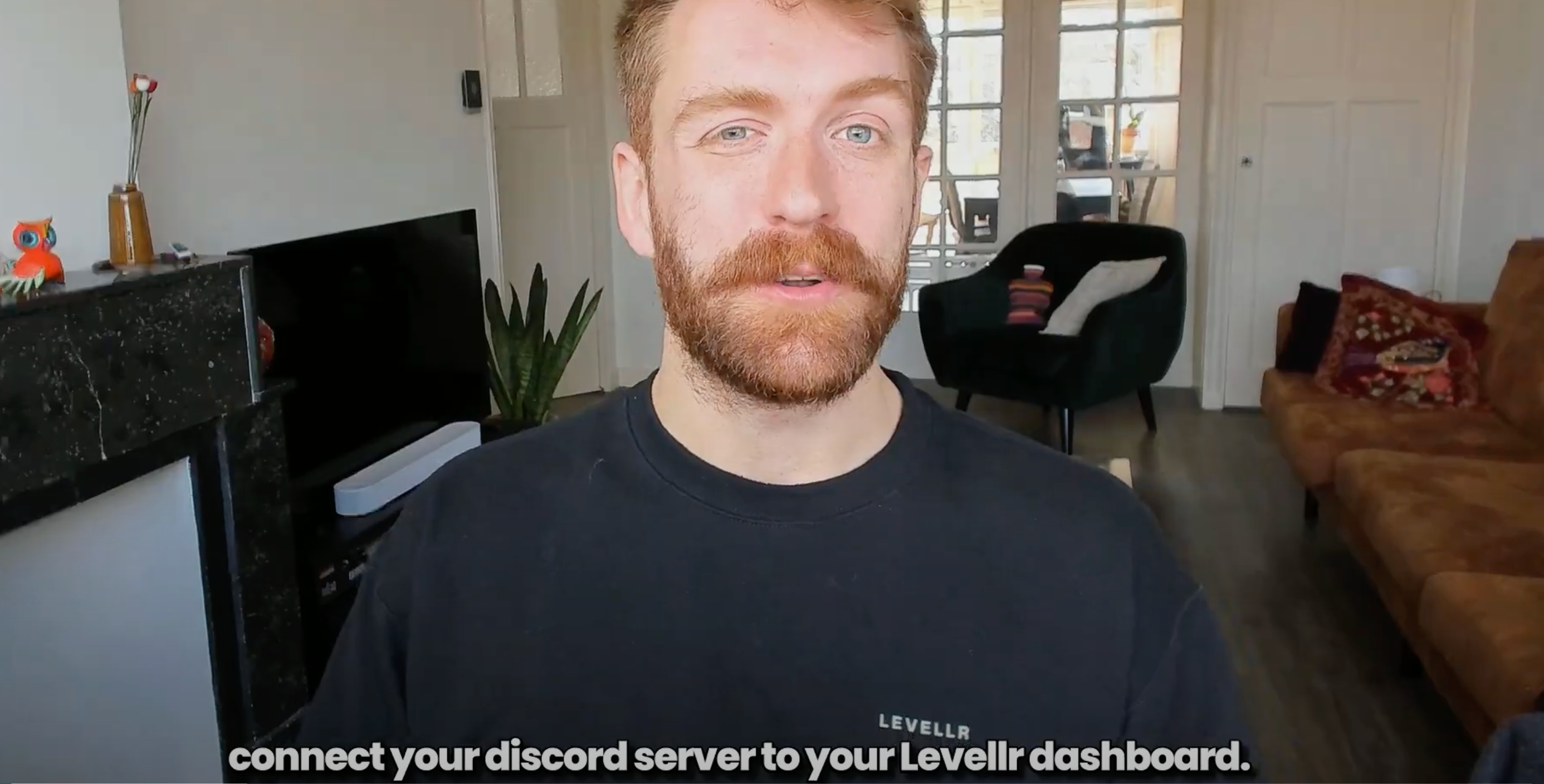 A man with short red hair and a beard speaks directly to the camera in a well-lit living room setting. He is wearing a black sweatshirt with the Levellr logo on the chest. Behind him, a TV, a fireplace, a plant, and a seating area with decorative pillows are visible. Subtitles at the bottom of the screen read, "connect your Discord server to your Levellr dashboard."