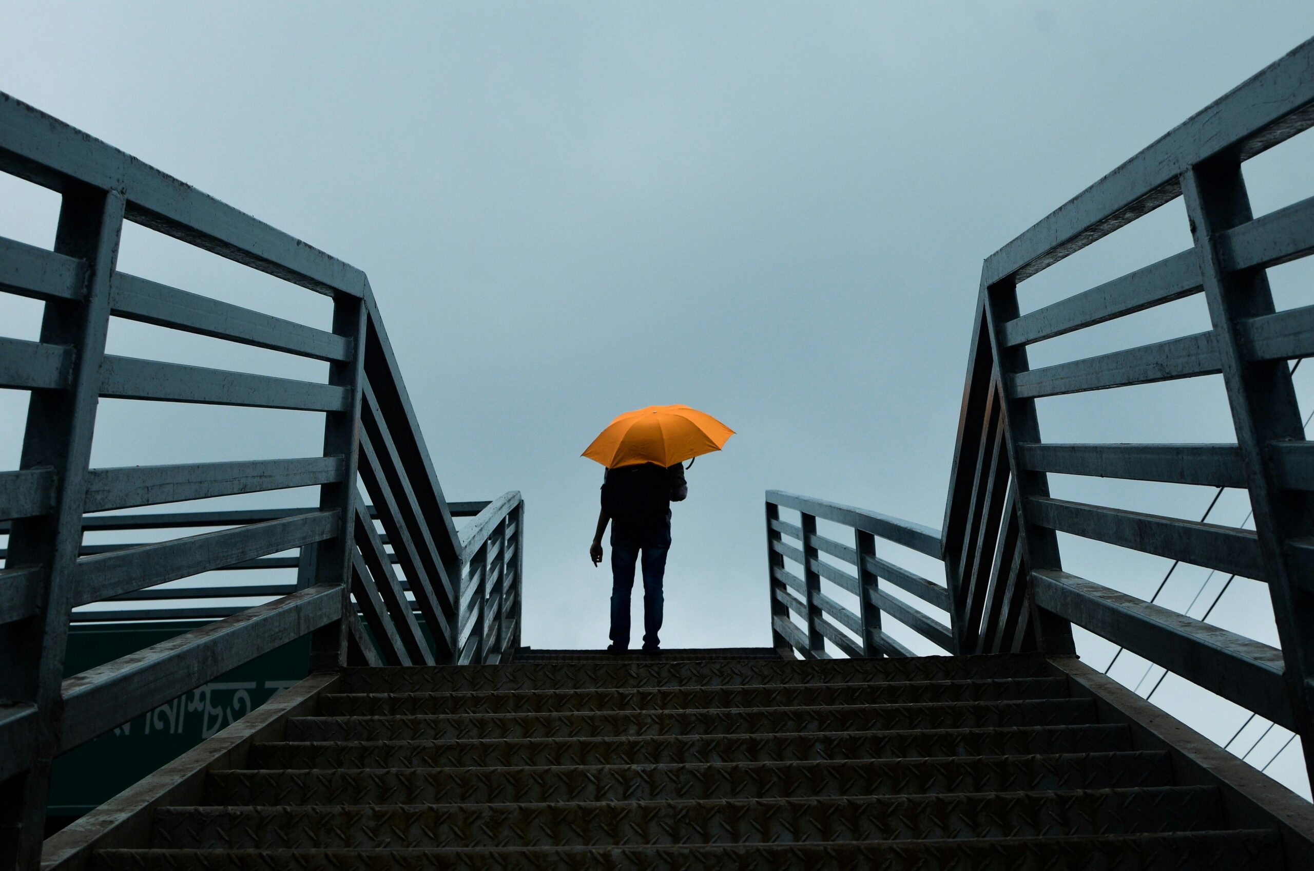 A person holding a bright orange umbrella stands at the top of a metal staircase on an overcast day. The stairway and railings frame the figure symmetrically, leading the viewer's eye towards the silhouette against the gray, cloudy sky. The contrast between the vivid umbrella and the muted surroundings emphasizes a sense of solitude and calm.